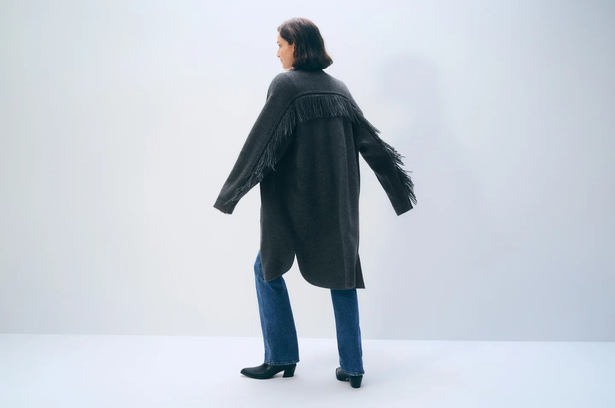 A woman is shown from the back wearing a dark charcoal gray fringed cardigan over blue jeans and black ankle boots, standing against a plain light gray studio background.