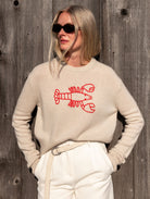 Woman wearing a beige sweater with a red lobster embroidered graphic and cream pants and a cream braided belt on a wooden background