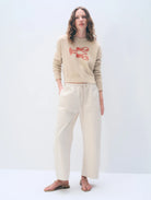 Woman wearing a beige sweater with red text and cream pants on a white background
