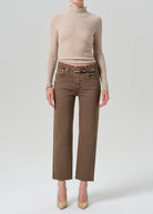 A model wearing a beige ribbed turtleneck sweater and brown wide-leg cropped jeans with a matching belt, paired with beige heels, against a light gray studio background.