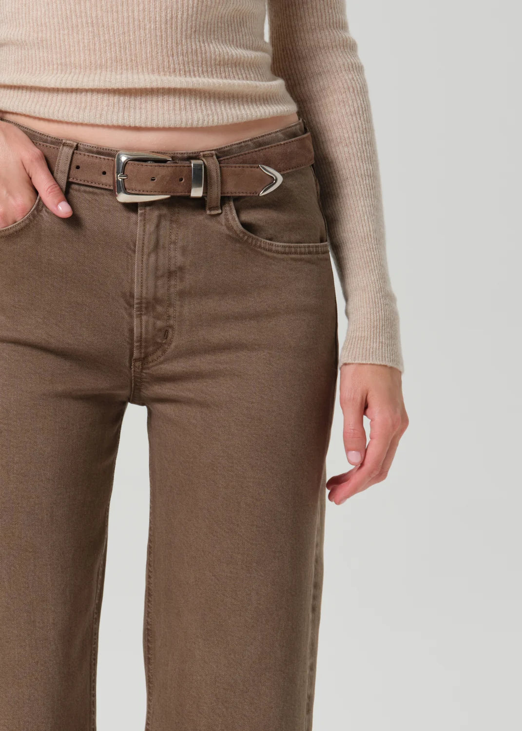 A close-up of a person wearing brown straight-leg jeans with a matching brown belt featuring a silver-toned metal tip, paired with a beige ribbed knit top against a light gray background.