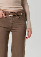 A close-up of a person wearing brown straight-leg jeans with a matching brown belt featuring a silver-toned metal tip, paired with a beige ribbed knit top against a light gray background.