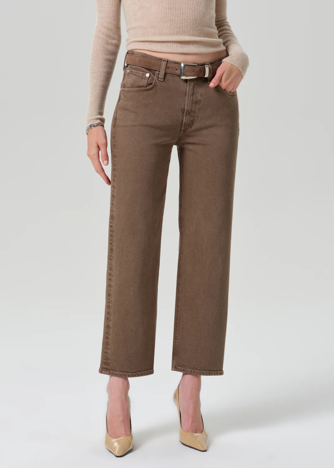 A model wearing brown cropped jeans with a matching belt, paired with a beige ribbed sweater and beige pointed-toe heels, photographed against a light gray studio background.