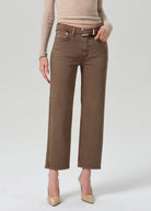 A model wearing brown cropped jeans with a matching belt, paired with a beige ribbed sweater and beige pointed-toe heels, photographed against a light gray studio background.