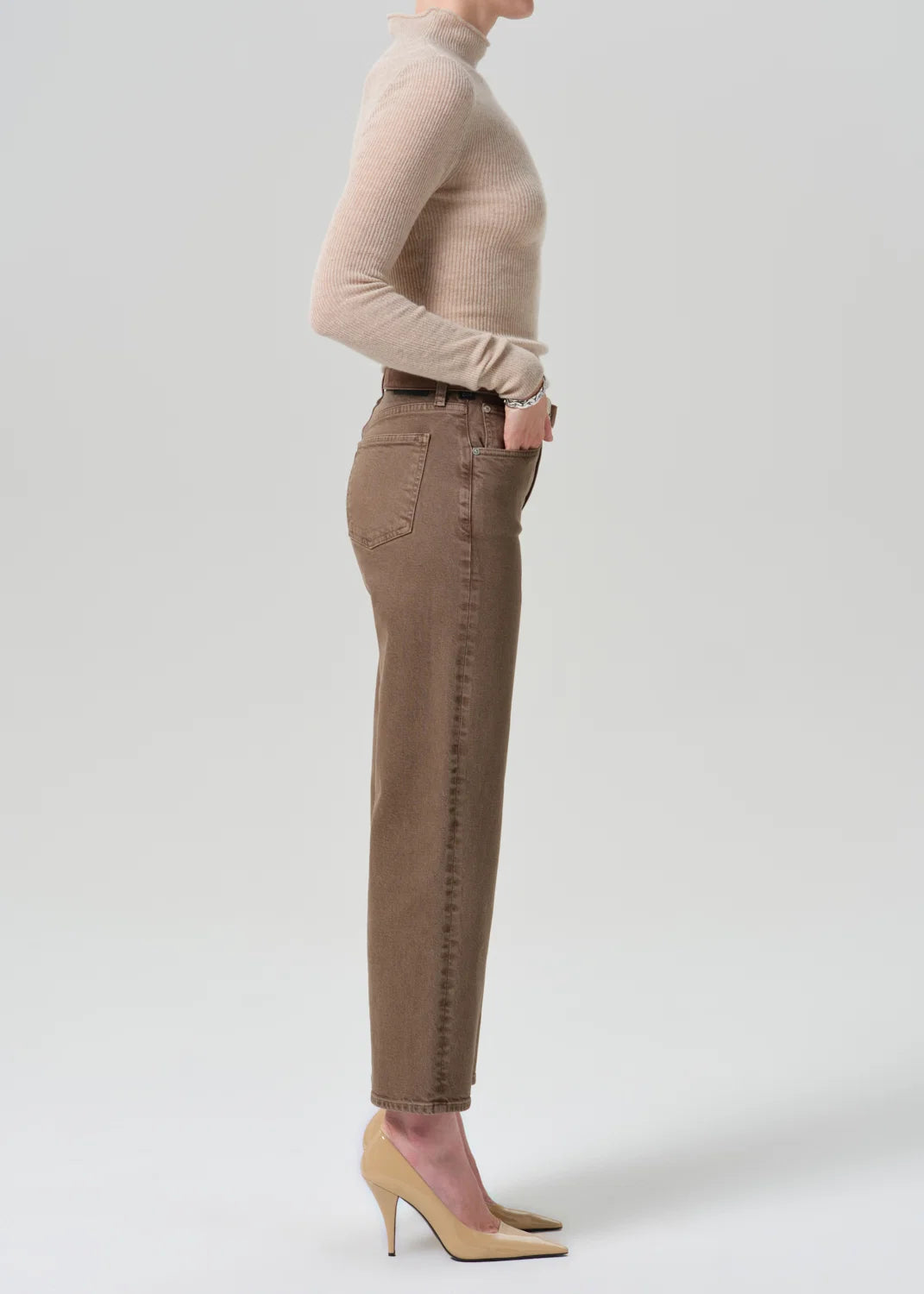 A side-profile studio shot of a model wearing brown ankle-length jeans with contrast stitching, a beige turtleneck sweater, and beige high heels against a light gray background.