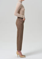 A side-profile studio shot of a model wearing brown ankle-length jeans with contrast stitching, a beige turtleneck sweater, and beige high heels against a light gray background.