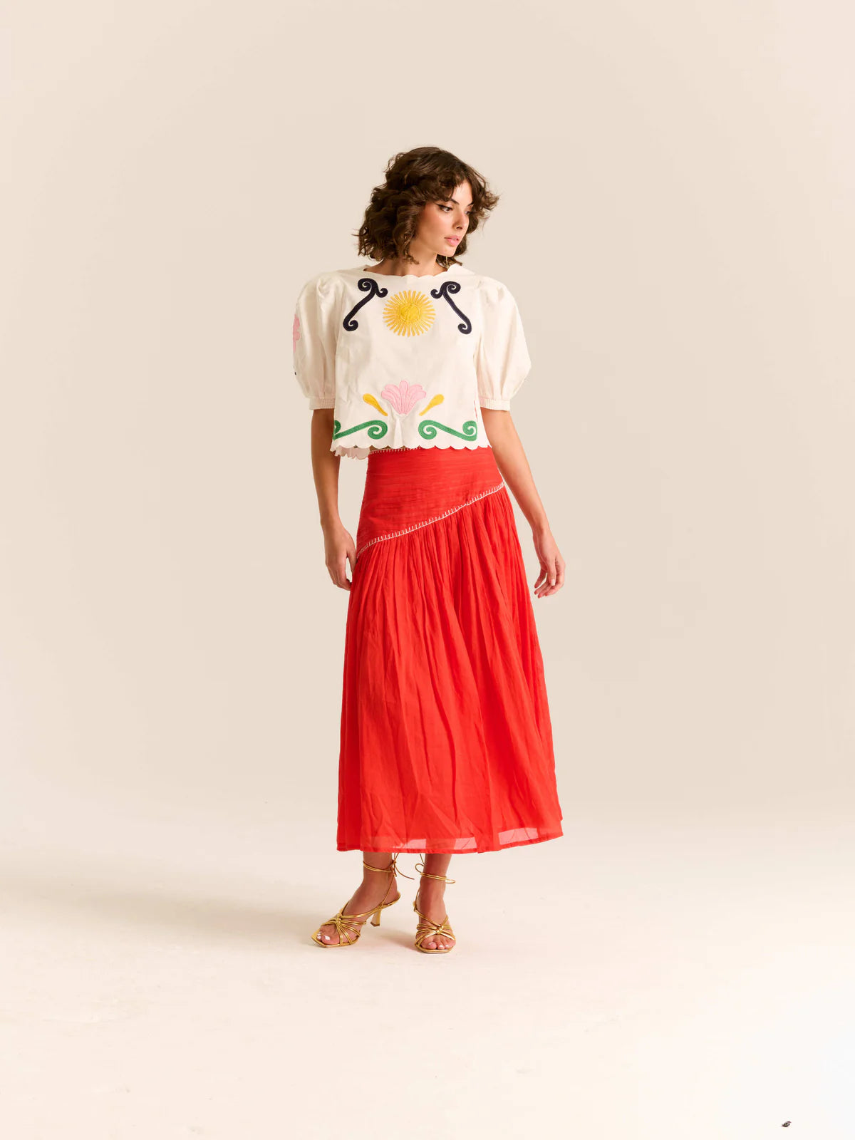 Woman wearing a white blouse with colorful patterns and a red skirt on a plain background