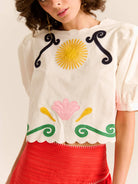 Woman wearing a white blouse with colorful embroidery and a red skirt on a plain background