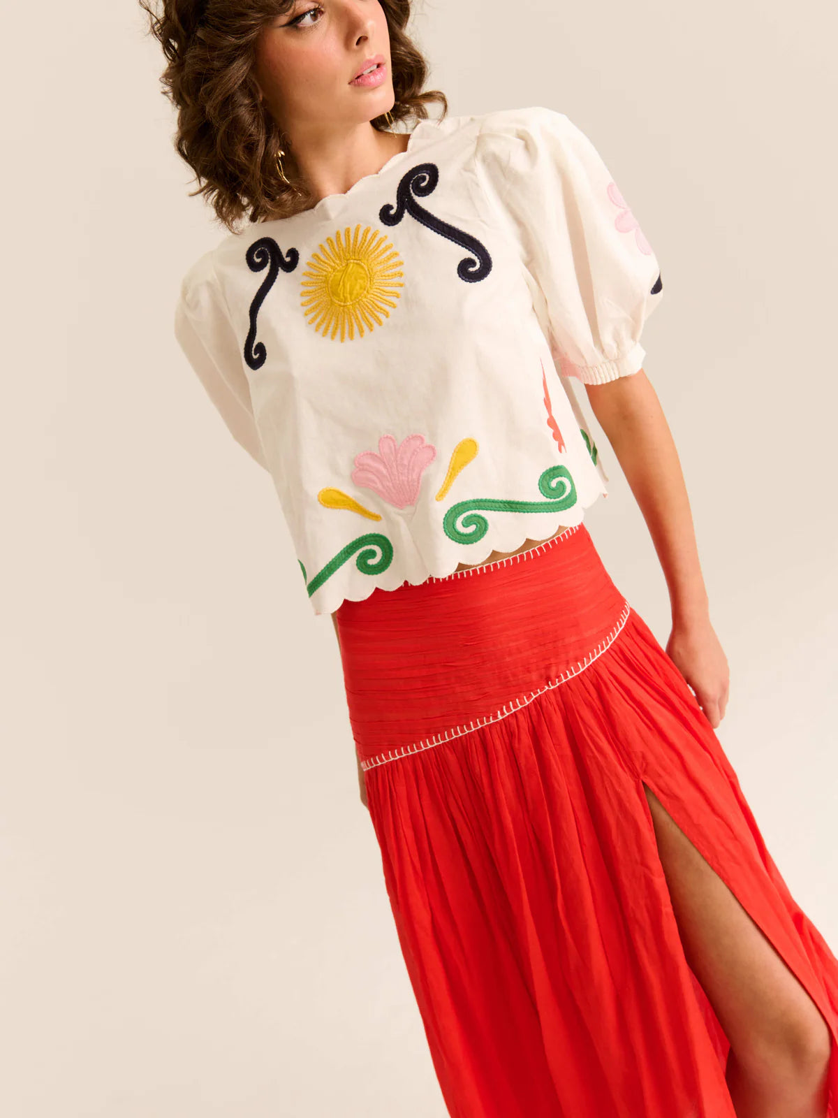 Woman wearing a white blouse with colorful designs and a red skirt on a beige background