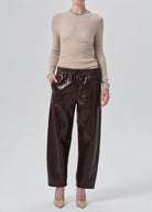 A model wearing a beige knit mock-neck sweater and dark brown wide-leg faux leather pants, standing against a light gray studio background.