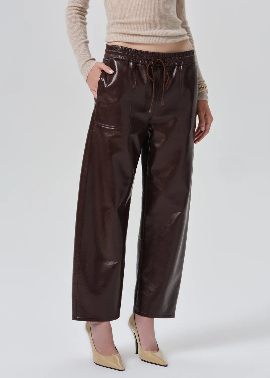 A model wearing shiny dark brown wide-leg cropped pants with an elastic drawstring waist, paired with a beige ribbed sweater and beige pointed-toe heels, against a light gray studio background.