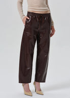 A model wearing shiny dark brown wide-leg cropped pants with an elastic drawstring waist, paired with a beige ribbed sweater and beige pointed-toe heels, against a light gray studio background.