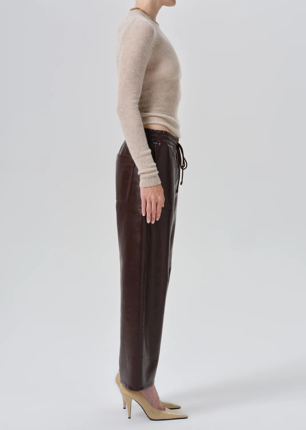 A side-profile studio shot of a model wearing dark brown faux-leather trousers with a drawstring waist, paired with a beige knit sweater and beige high heels, against a light gray background.