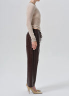 A side-profile studio shot of a model wearing dark brown faux-leather trousers with a drawstring waist, paired with a beige knit sweater and beige high heels, against a light gray background.