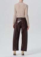 A model shown from the back wearing a beige knit sweater and dark brown wide-leg faux leather trousers, paired with beige high heels, against a light gray studio background.