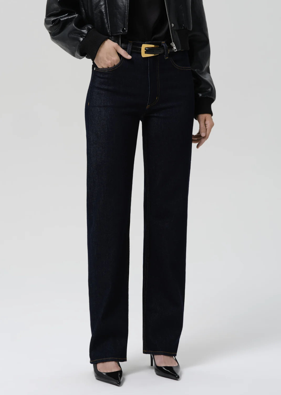 A model wearing high-waisted, straight-leg dark wash jeans with a gold-tone belt buckle, styled with a black leather jacket, black top, and black pointed-toe heels against a light gray studio background.