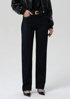 A model wearing high-waisted, straight-leg dark wash jeans with a gold-tone belt buckle, styled with a black leather jacket, black top, and black pointed-toe heels against a light gray studio background.