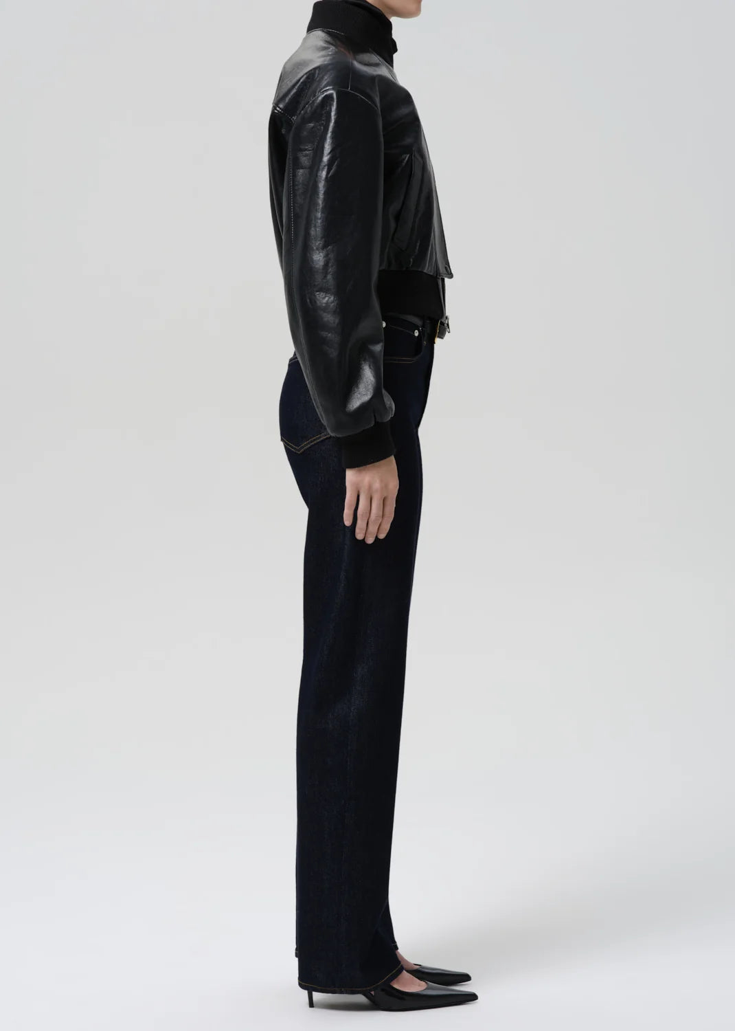 A model shown from the side wearing a black leather cropped jacket, black turtleneck, black wide-leg trousers, and black pointed-toe flats against a light gray studio background.