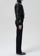 A model shown from the side wearing a black leather cropped jacket, black turtleneck, black wide-leg trousers, and black pointed-toe flats against a light gray studio background.