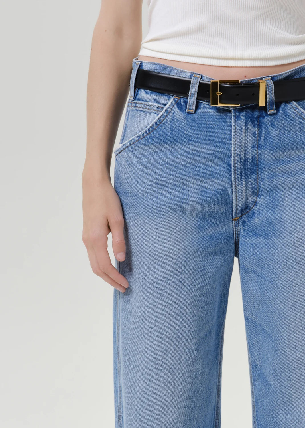 Close-up of a person wearing light blue jeans with a black leather belt featuring gold-tone hardware, paired with a white ribbed top against a plain light gray background.
