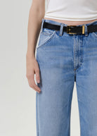 Close-up of a person wearing light blue jeans with a black leather belt featuring gold-tone hardware, paired with a white ribbed top against a plain light gray background.