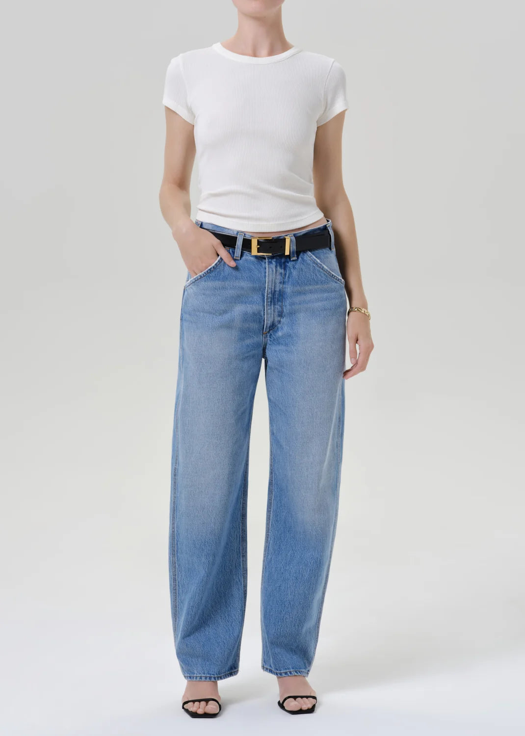 A model wearing a white short-sleeve crewneck t-shirt, light-wash blue wide-leg jeans with a black belt, and black sandals, standing against a light gray studio background.