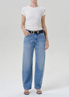 A model wearing a white short-sleeve crewneck t-shirt, light-wash blue wide-leg jeans with a black belt, and black sandals, standing against a light gray studio background.