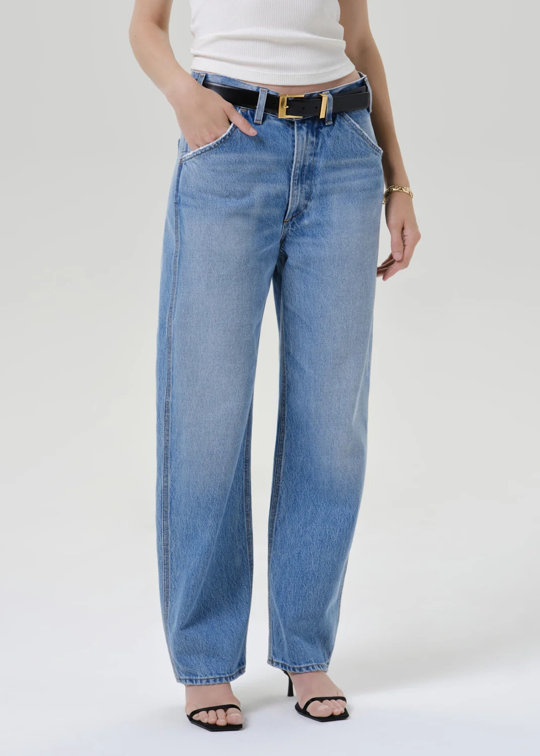 A model wearing a white short-sleeve crewneck t-shirt, light-wash blue wide-leg jeans with a black belt, and black sandals, standing against a light gray studio background.
