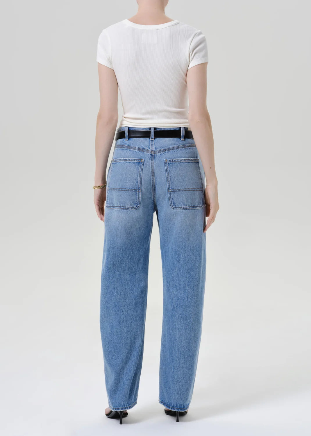 A model shown from the back wearing light-wash blue wide-leg jeans, a white short-sleeve t-shirt, and black heels against a plain light gray studio background.