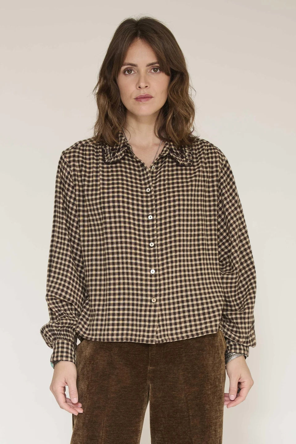 A woman wearing a brown and beige gingham long-sleeve button-up shirt with brown corduroy pants, standing against a plain light beige studio background.