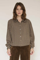 A woman wearing a brown and beige gingham long-sleeve button-up shirt with brown corduroy pants, standing against a plain light beige studio background.