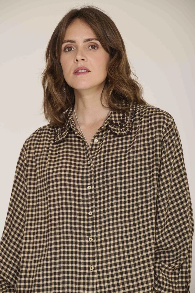 A woman with shoulder-length brown hair wearing a brown and beige gingham button-up shirt, photographed against a plain light beige studio background.