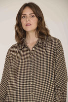 A woman with shoulder-length brown hair wearing a brown and beige gingham button-up shirt, photographed against a plain light beige studio background.