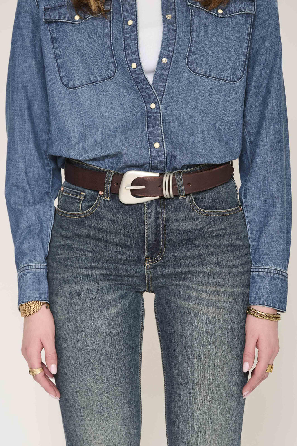 A person wearing a cropped blue denim shirt tucked into high-waisted blue jeans, styled with a dark brown belt featuring a silver-toned buckle.
