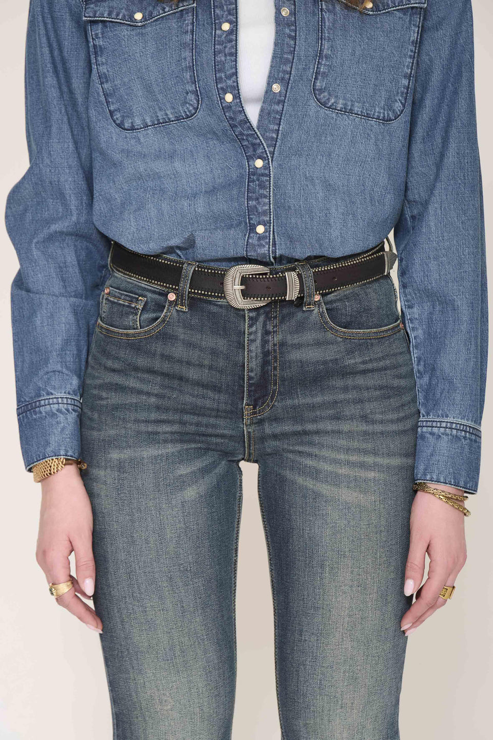 A person wearing a blue denim button-up shirt tucked into blue jeans, with a dark brown belt featuring silver-toned studs and a silver buckle. The photo is cropped from the chest down against a plain light background.