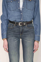 A person wearing a blue denim button-up shirt tucked into blue jeans, with a dark brown belt featuring silver-toned studs and a silver buckle. The photo is cropped from the chest down against a plain light background.