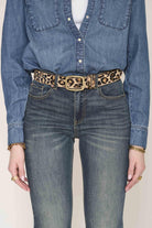 A close-up of a person wearing a leopard-print belt with a gold-tone rectangular buckle, styled with a blue denim shirt and blue jeans against a light beige background.