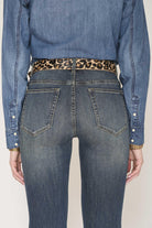 Rear view of a person wearing blue denim jeans with a leopard-print belt, a blue denim shirt, and a gold bracelet, against a light beige background.