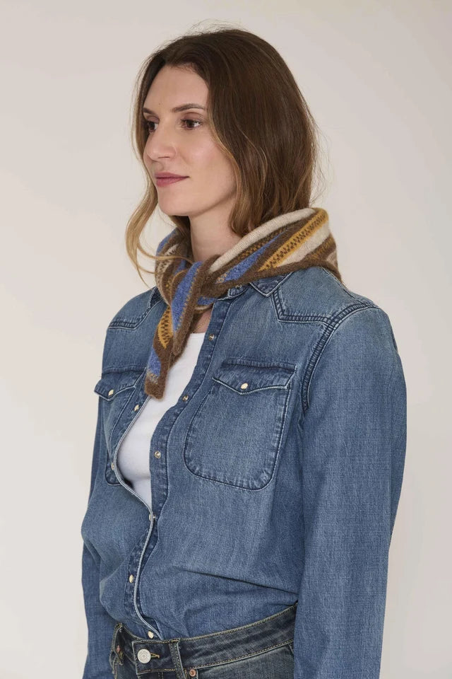 A woman wearing a blue denim shirt jacket over a white top, styled with a brown and blue patterned knit scarf, photographed against a plain light background.