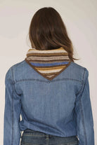A woman viewed from behind wearing a blue denim jacket over a striped knit cowl neck scarf in brown, beige, and blue tones.