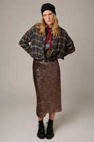 A model stands against a plain light gray studio background wearing a black beanie, a dark plaid button-up shirt, a red necktie, a colorful graphic top, a brown sequined midi skirt, and black lace-up ankle boots.