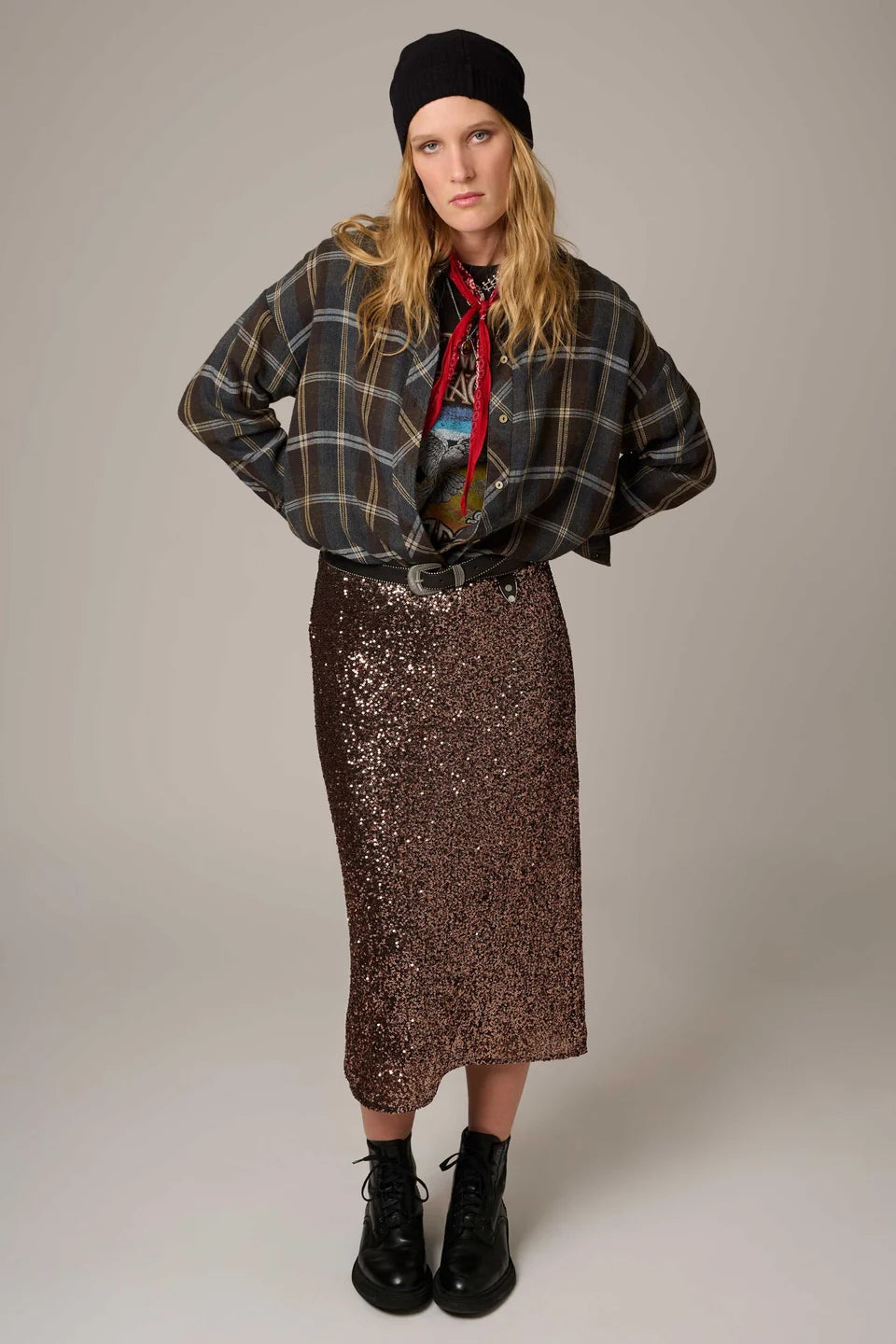 A model stands against a plain light gray studio background wearing a black beanie, a dark plaid button-up shirt, a red necktie, a colorful graphic top, a brown sequined midi skirt, and black lace-up ankle boots.