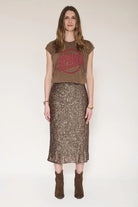 A woman stands against a plain light beige studio background wearing a brown cropped T-shirt with a faded red circular graphic and a brown sequined midi skirt with a ruffled hem, paired with brown ankle boots.