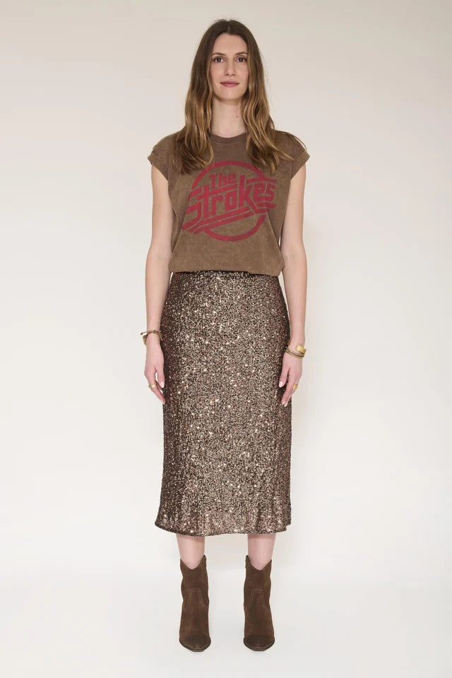 A woman stands against a plain light beige studio background wearing a brown cropped T-shirt with a faded red circular graphic and a brown sequined midi skirt with a ruffled hem, paired with brown ankle boots.