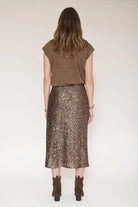 A woman shown from the back wearing a brown sleeveless top and a shimmering bronze sequined midi skirt, paired with brown ankle boots, against a plain light background.