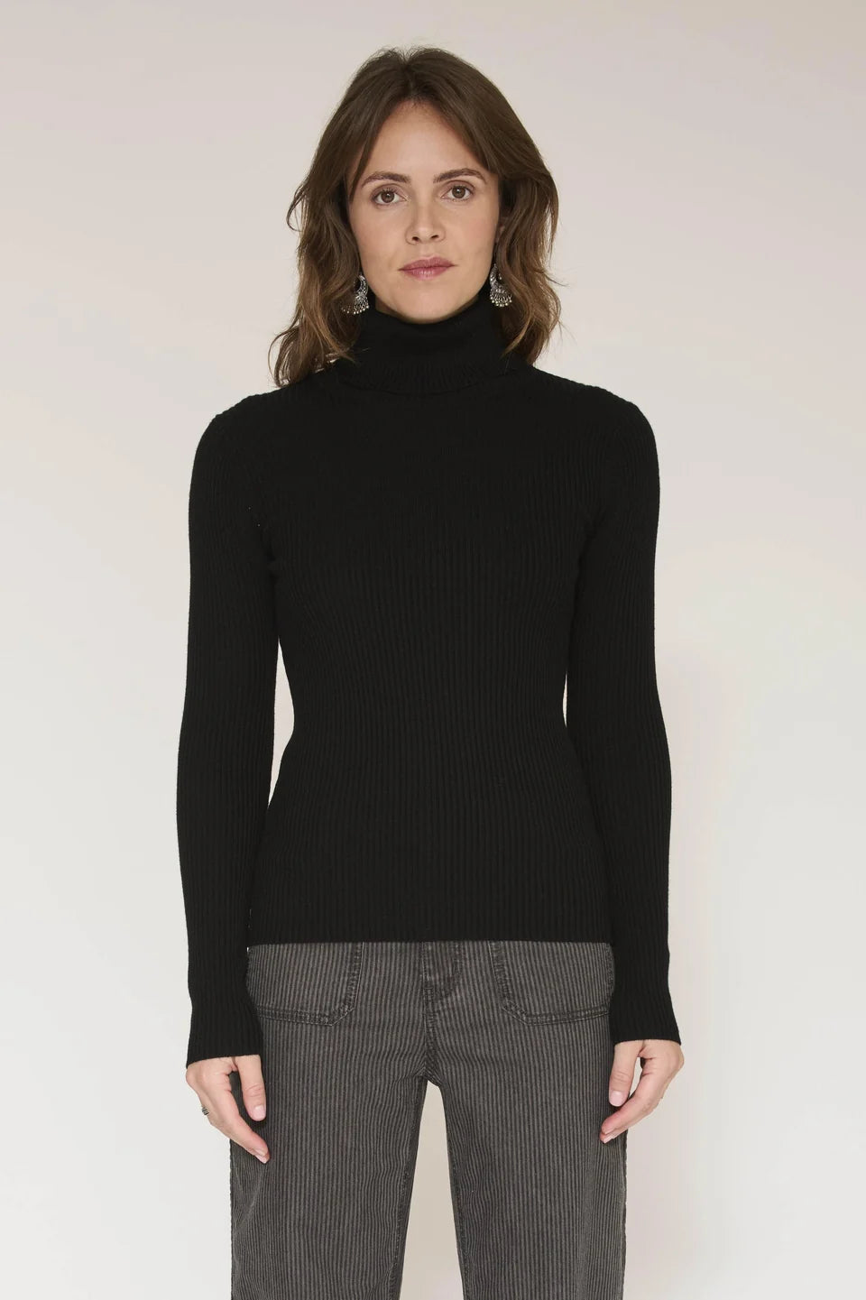 A woman modeling a black ribbed turtleneck sweater with long sleeves, paired with gray pinstriped trousers, against a plain light background.