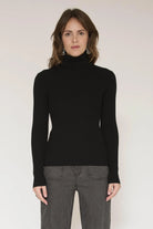 A woman modeling a black ribbed turtleneck sweater with long sleeves, paired with gray pinstriped trousers, against a plain light background.