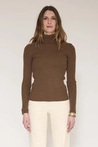 A woman modeling a brown ribbed knit mock neck sweater with cream pants, standing against a plain light background.