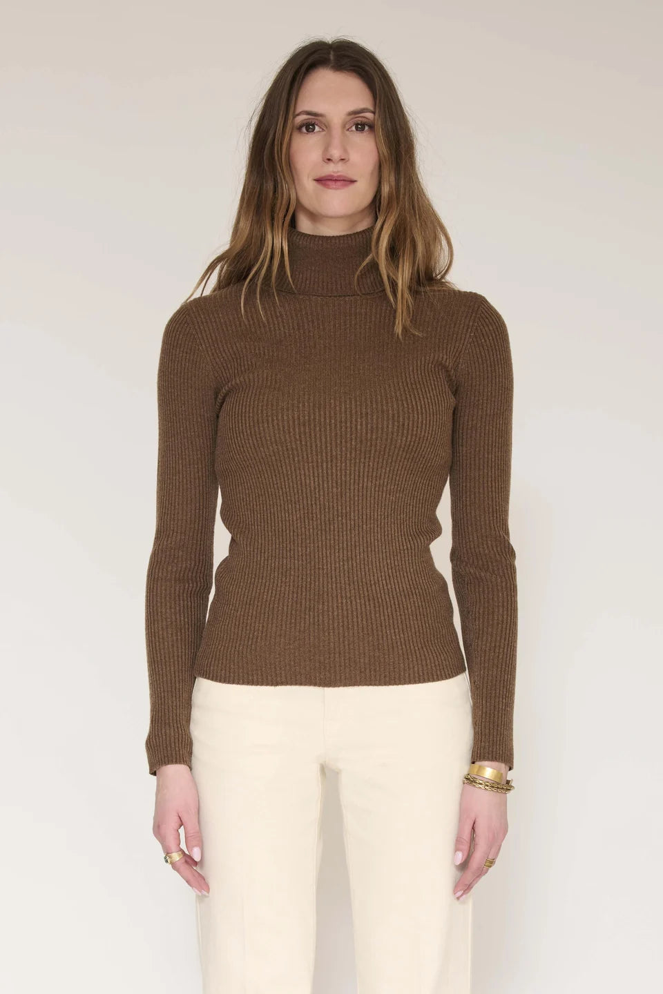 A woman modeling a brown ribbed knit mock neck sweater with cream pants, standing against a plain light background.