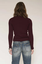 A woman shown from the back wearing a ribbed long-sleeve sweater in deep burgundy and blue jeans, against a plain light beige studio background.